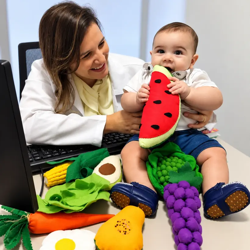 Lígia de jaleco branco no consultório, segurando um bebê que morde uma melancia de feltro, com frutas e legumes de feltro sobre a mesa