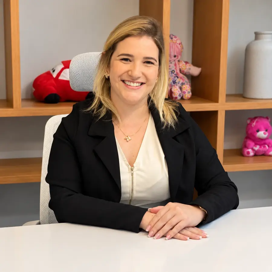 Lígia Valença sorrindo à mesa do consultório, de blazer preto, com pelúcias na estante ao fundo